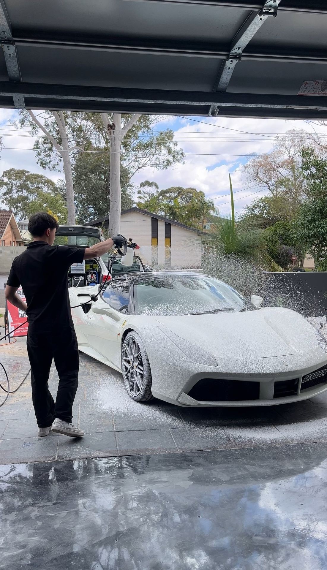 Person washing a white sports car with foam in a driveway, surrounded by trees and houses.