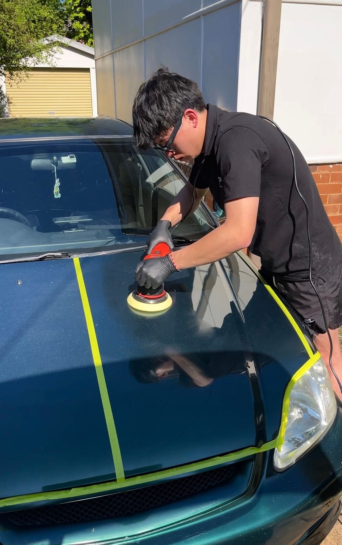 Person in black attire using a polisher on a car hood with taped sections.