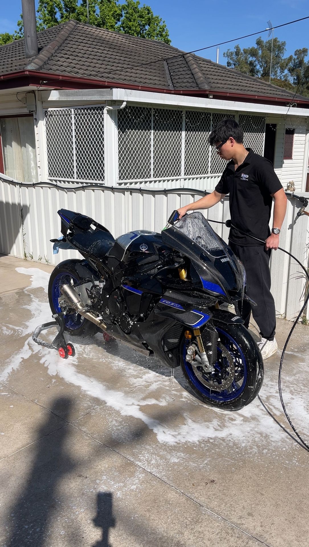 Person washing a black and blue motorcycle with soap near a white house on a sunny day.